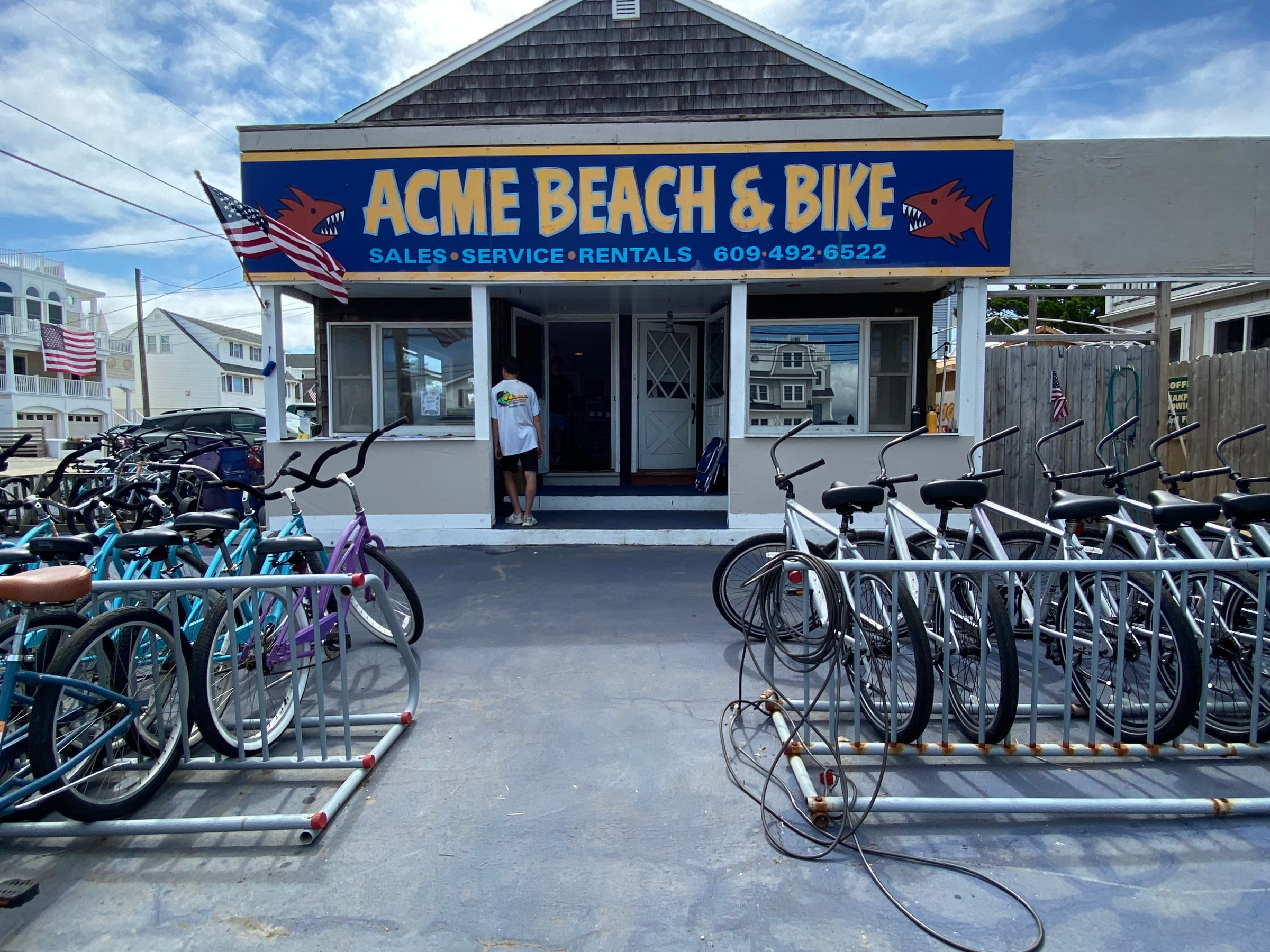 Bicycle shop with bikes outside and a sign reading 'ACME BEACH & BIKE' under a cloudy sky.