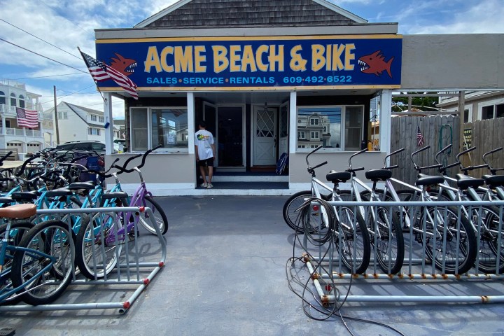 Bicycle shop with bikes outside and a sign reading 'ACME BEACH & BIKE' under a cloudy sky.