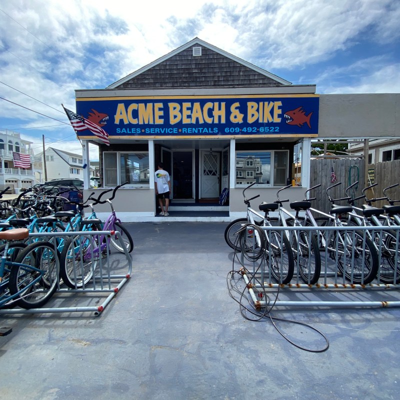 Bicycle shop with bikes outside and a sign reading 'ACME BEACH & BIKE' under a cloudy sky.
