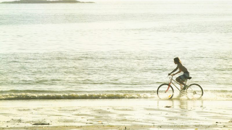 Person rides a bicycle along the beach shore with an island in the background.
