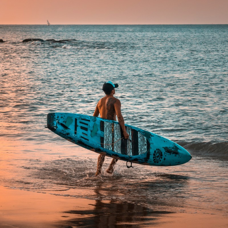Person carrying a surfboard into the ocean during sunset.