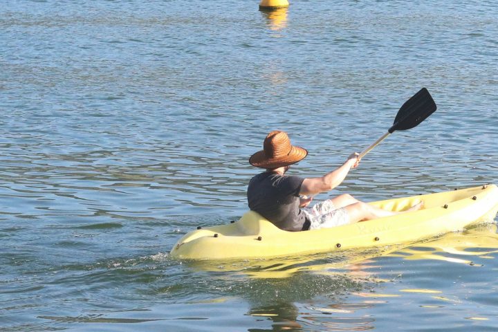 Person in a hat kayaking on calm water with yellow buoys in the distance.
