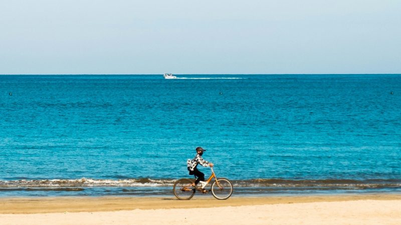 Person riding a bicycle on a beach with ocean and clear blue sky.
