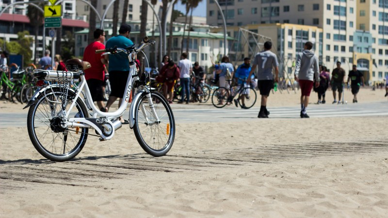 White bike parked on sandy beach with people and bikes in background.